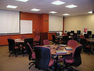 Lab room with desks along walls, with computer stations and black chairs at each station. A faculty member using one station.